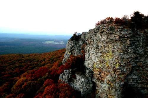 A rocky cliff overlooks a vibrant autumn landscape filled with colorful trees and distant hills.