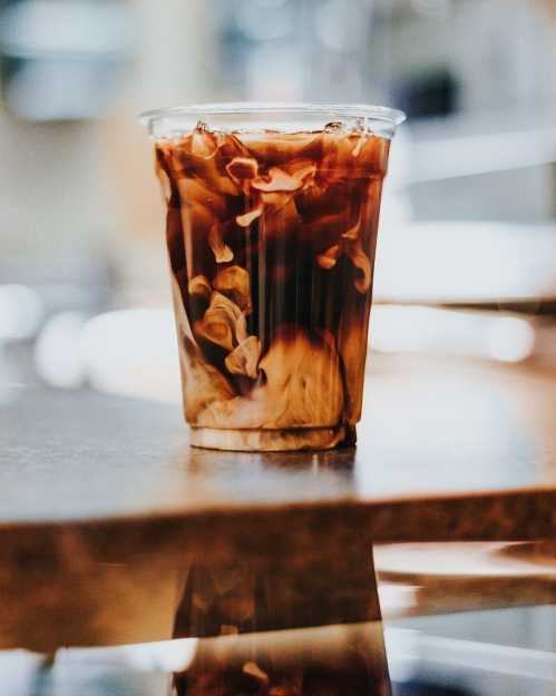 A clear cup filled with iced coffee, swirling with cream, sits on a wooden table.