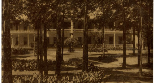 Historic building surrounded by trees and gardens, featuring a porch and multiple balconies.