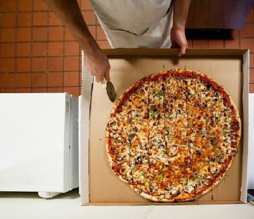 A person using a pizza cutter to slice a large pizza in a cardboard box.