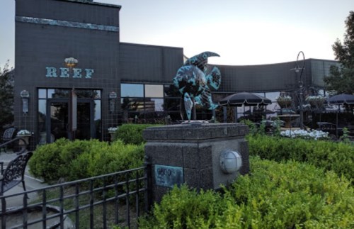 A restaurant named "Reef" with a sculpture in front, surrounded by greenery and outdoor seating.