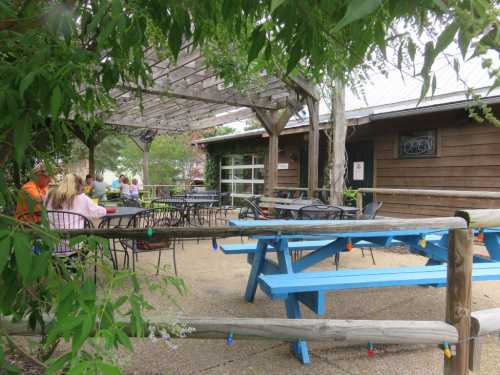 Outdoor dining area with blue picnic tables and a wooden pergola, surrounded by greenery and a rustic building.