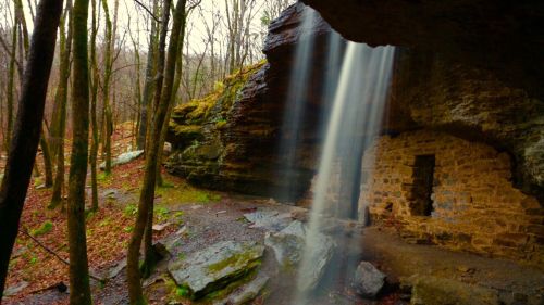 A serene waterfall cascades from a rocky overhang in a forest, surrounded by trees and moss-covered stones.