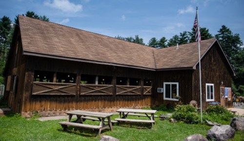 A wooden building with a flag, surrounded by trees and picnic tables in a sunny outdoor setting.