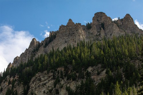 A rugged mountain peak rises above a dense forest under a blue sky with scattered clouds.