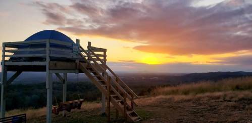 A wooden platform with a blue dome structure overlooks a sunset, surrounded by rolling hills and grassy fields.