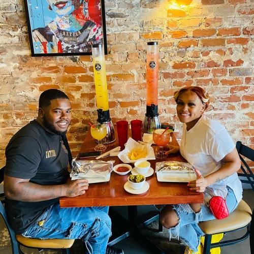 A couple sits at a table with food and colorful drinks in a cozy restaurant with a brick wall and artwork.