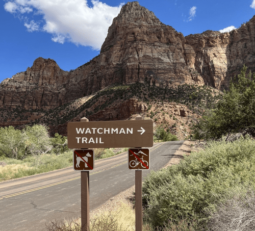 Sign for Watchman Trail with symbols for hiking and biking, set against a backdrop of rocky mountains and blue sky.