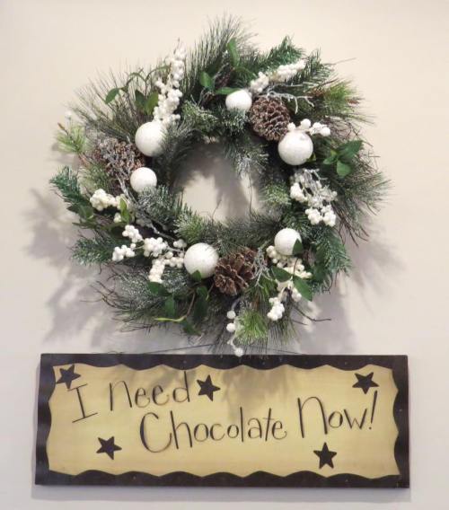 A festive wreath with pinecones and white ornaments, accompanied by a sign that says, "I Need Chocolate Now!"