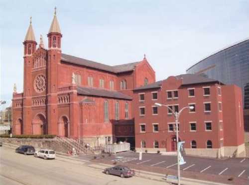 Historic red brick church with twin towers, adjacent to a modern building, set along a city street.