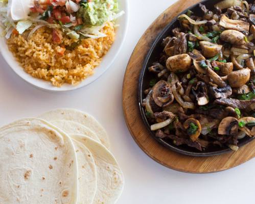 A sizzling platter of beef fajitas with mushrooms and onions, served with rice, salad, and tortillas on the side.