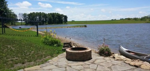 A serene lakeside view with a stone fire pit, canoe, hammock, and lush greenery under a clear blue sky.