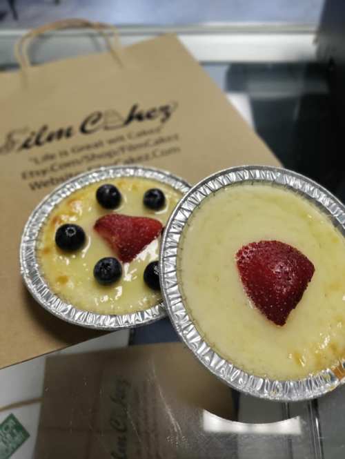 Two small dessert cups with creamy custard topped with a strawberry and blueberries, next to a brown paper bag.