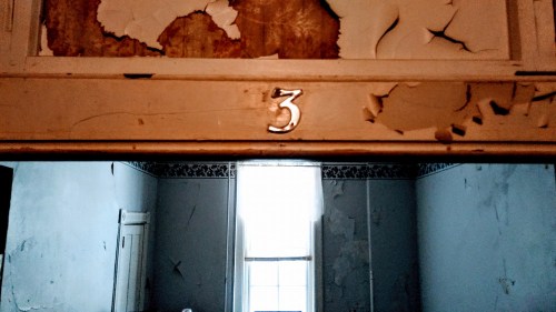 A weathered wall with peeling paint features the number 3 above a doorway, leading to a bright window in the background.