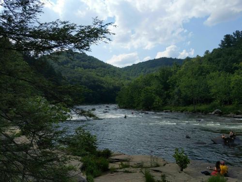 A serene river flows through a lush green landscape, surrounded by mountains and dotted with people enjoying the outdoors.