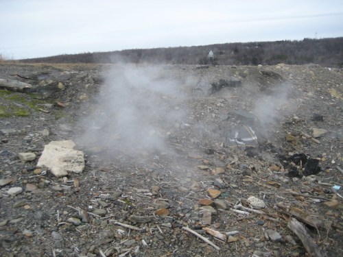 Steam rising from rocky terrain, with scattered stones and a cloudy sky in the background.