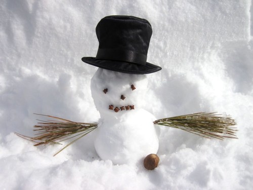 A snowman with a black hat, twig arms, and a face made of small stones, sitting in a snowy landscape.