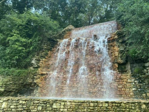 A cascading waterfall flows over a rocky cliff, surrounded by lush greenery and trees.