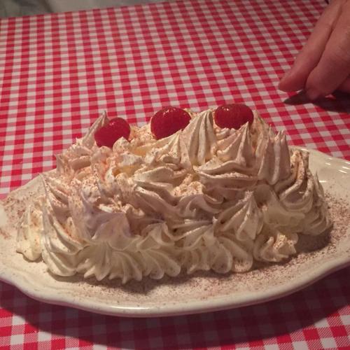 A dessert topped with whipped cream and raspberries, served on a white plate, on a red and white checkered tablecloth.