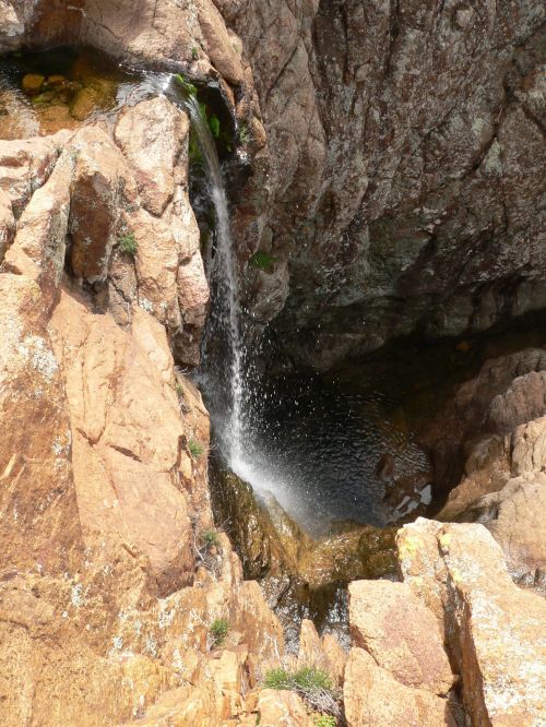 A small waterfall cascading over rocky terrain into a clear pool below, surrounded by rugged stone formations.