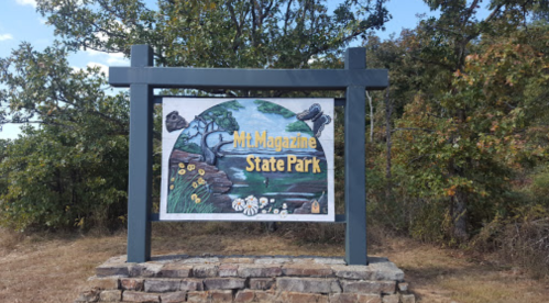 Sign for Mt. Magazine State Park featuring a colorful design and surrounded by trees and grass.
