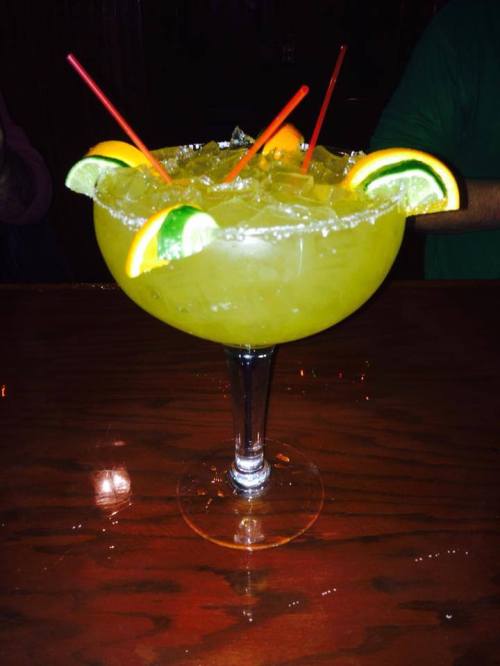 A large margarita glass filled with a green drink, ice, and garnished with orange and lime slices, topped with red straws.