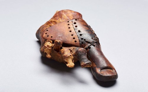 A carved wooden artifact with holes and decorative stitching, resting on a textured surface.