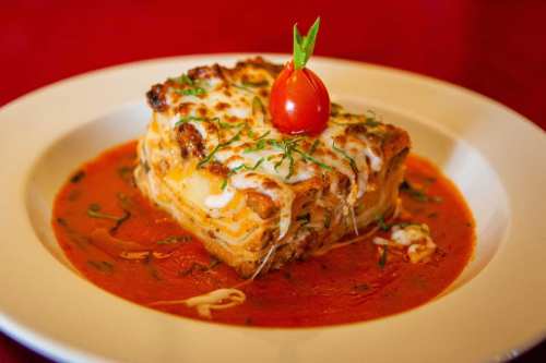 A slice of lasagna topped with a cherry tomato, served in a rich tomato sauce with herbs.