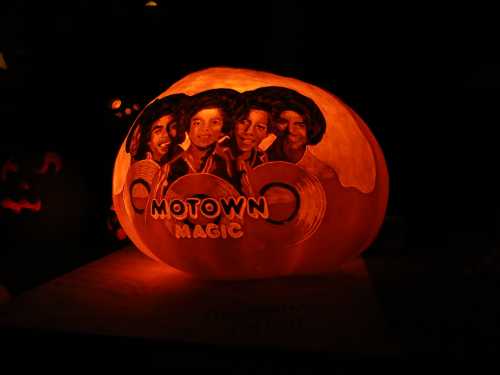 A carved pumpkin glowing orange, featuring the words "Motown Magic" and portraits of four musicians.