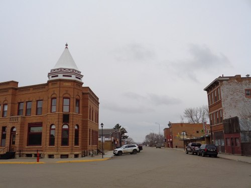 A quiet street scene featuring historic brick buildings and a cloudy sky. Vehicles are parked along the road.