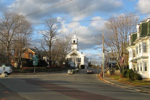 A quaint town scene featuring a white church, trees, and houses under a partly cloudy sky.