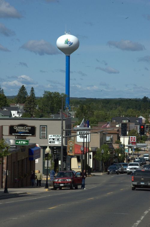 A small town street with shops, a water tower in the background, and a clear blue sky with scattered clouds.