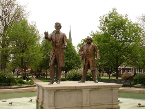 Statue of Abraham Lincoln and another figure on a pedestal in a park, surrounded by greenery and a fountain.