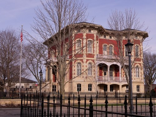 Historic building with red brick and cream stone, surrounded by trees and a black iron fence, under a clear sky.