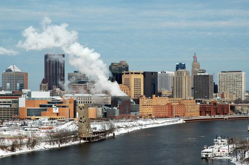 A winter cityscape featuring buildings along a river, with smoke rising and snow covering the ground.