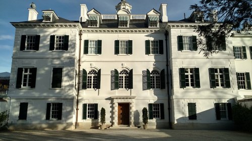A large, elegant white mansion with black shutters and a central entrance, surrounded by greenery and a clear sky.