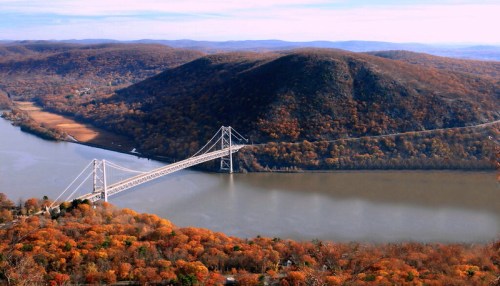 Aerial view of a bridge spanning a river, surrounded by autumn foliage and rolling hills.