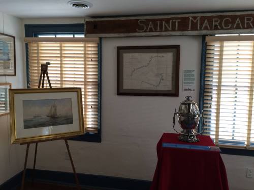 A room with wooden blinds, featuring a framed painting of a sailboat, a map, and a decorative silver piece on a table.