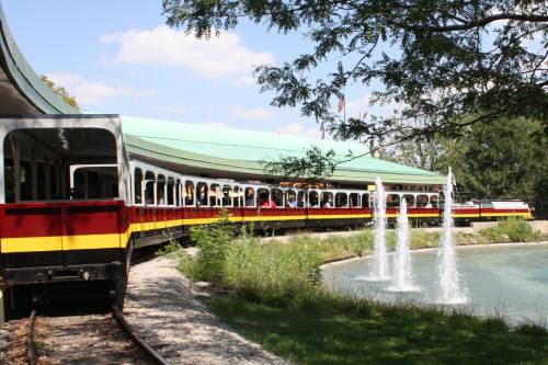 A colorful train curves around a pond with fountains, surrounded by greenery and a blue sky.