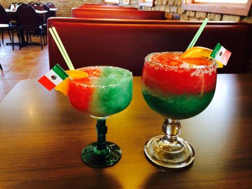 Two colorful frozen cocktails in decorative glasses, featuring red, green, and orange layers, with small Mexican flags.