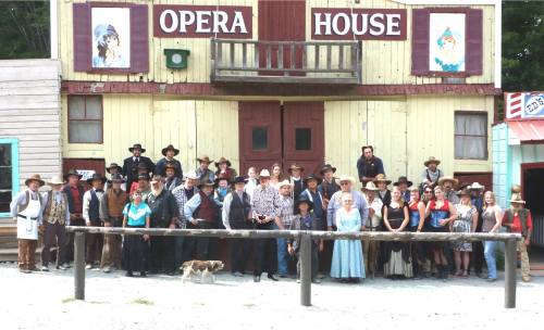 A large group of people in Western attire poses in front of a colorful opera house building. A dog is also present.