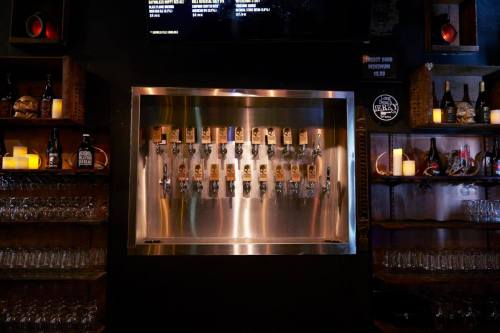 A sleek bar with a metal tap wall featuring multiple beer taps and shelves stocked with bottles and glasses.
