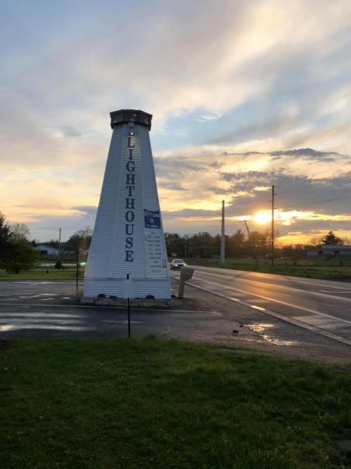 A lighthouse-shaped sign stands by the road, with a colorful sunset in the background and clouds in the sky.
