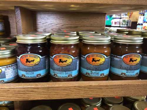 Jars of various sauces and butters on a wooden shelf, labeled with colorful designs and text.