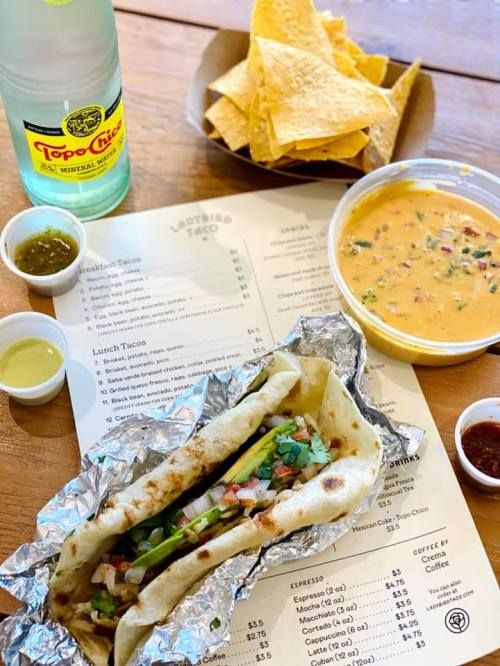 A taco with toppings, a bowl of cheese dip, tortilla chips, and a bottle of sparkling water on a wooden table.