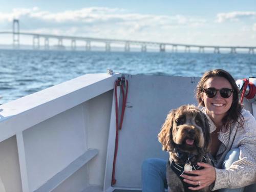 A woman in sunglasses sits on a boat with a dog, with a bridge visible in the background and water all around.