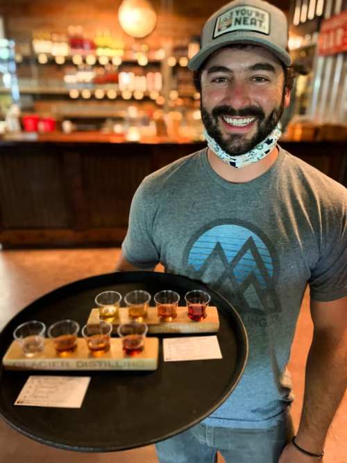 A smiling man in a gray t-shirt holds a tray with eight small glasses of different colored drinks at a distillery.
