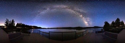 A panoramic view of a starry night sky over a calm lake, with silhouettes of trees and a wooden deck in the foreground.