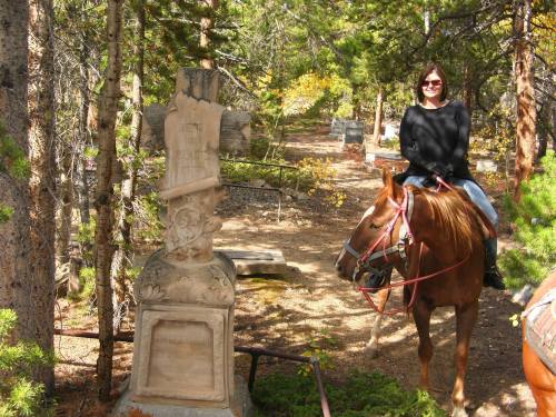 A person on a horse next to a stone cross in a wooded area, surrounded by trees and autumn foliage.