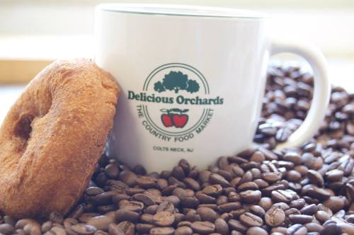 A white mug with a logo next to a donut, surrounded by coffee beans on a wooden surface.